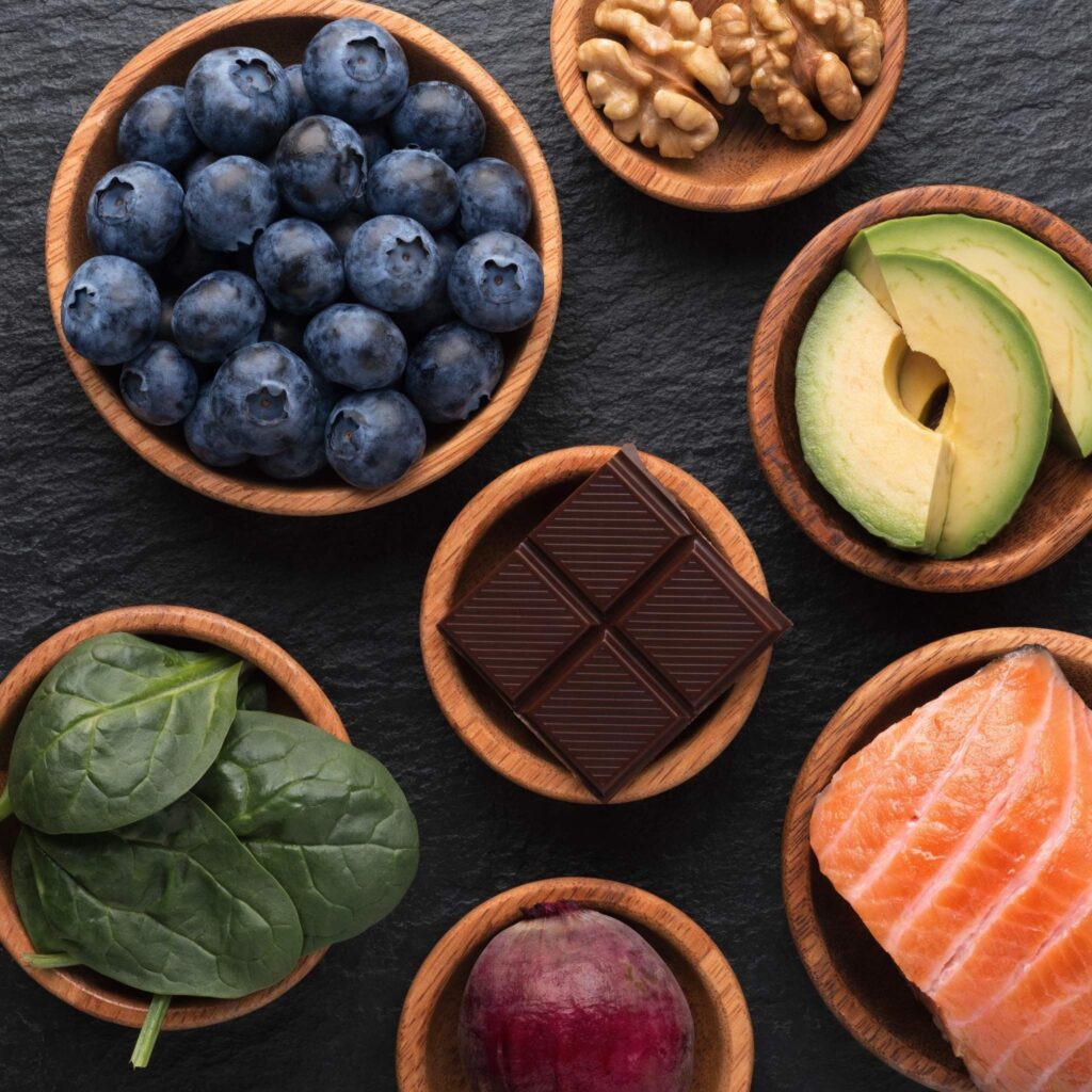 Seven brain-boosting foods for seniors including blueberries, walnuts, avocado, dark chocolate, spinach, beets, and salmon arranged in wooden bowls on a slate surface